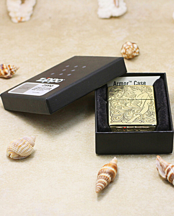 Black Zippo lighter with gold floral engraving in an open black box on a sandy surface with seashells nearby.