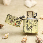 Gold engraved Zippo-style lighter opened on a sandy beach, with seashells around it.