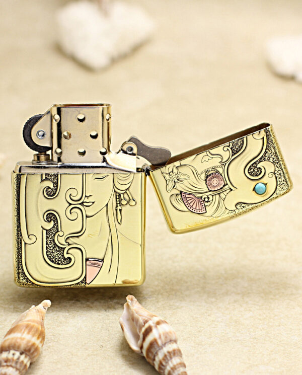 Close-up of a gold, engraved Zippo lighter on sand with seashells nearby, the lid open.
