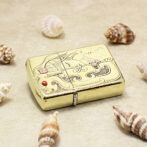 Gold decorative lighter with engraved wave pattern resting on sand, surrounded by seashells (beach theme).