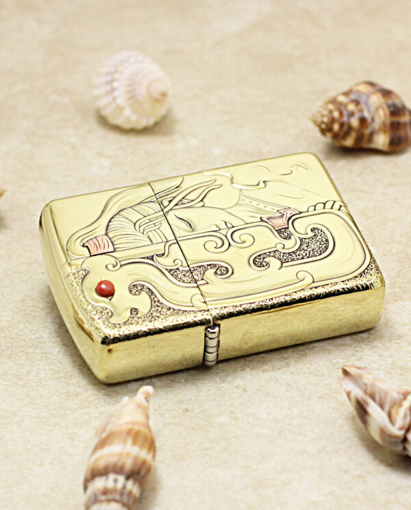 Gold decorative lighter with engraved wave pattern resting on sand, surrounded by seashells (beach theme).