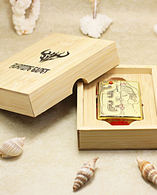 Gold Zippo lighter with decorative engravings in a light wooden gift box, surrounded by seashells at the beach.