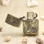 Gold brass Zippo-style lighter with an ornate dragon and floral engraving, open on a sandy surface with seashells nearby