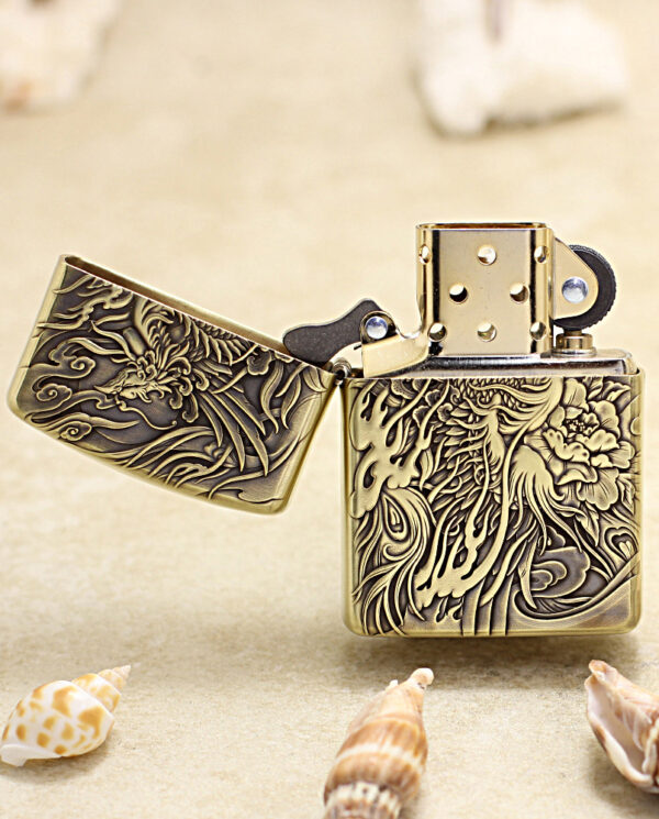 KZ229-02 Gold brass Zippo-style lighter with an ornate dragon and floral engraving, open on a sandy surface with seashells nearby