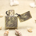 Brass Zippo-style lighter with a dragon engraving, open lid, on a sandy beach with seashells nearby.