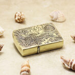 Brass lighter with intricate dragon engraving, placed on sandy surface among seashells.