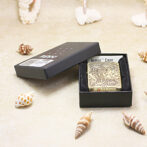 Engraved brass Zippo lighter in Armor Case inside a black presentation box on a sandy surface with seashells nearby.