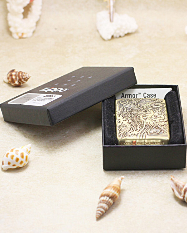 KZ229-06 Engraved brass Zippo lighter in Armor Case inside a black presentation box on a sandy surface with seashells nearby.