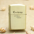Gold-toned Zippo-style lighter with engraving, standing on sand among seashells (0772/1000).