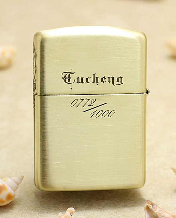 Gold-toned Zippo-style lighter with engraving, standing on sand among seashells (0772/1000).