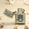 Gold Zippo lighter with Freemason square-and-compass emblem and open lid, set on a sandy beach with seashells nearby.