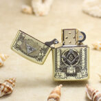 Gold Zippo lighter with Freemason square-and-compass emblem and open lid, set on a sandy beach with seashells nearby.