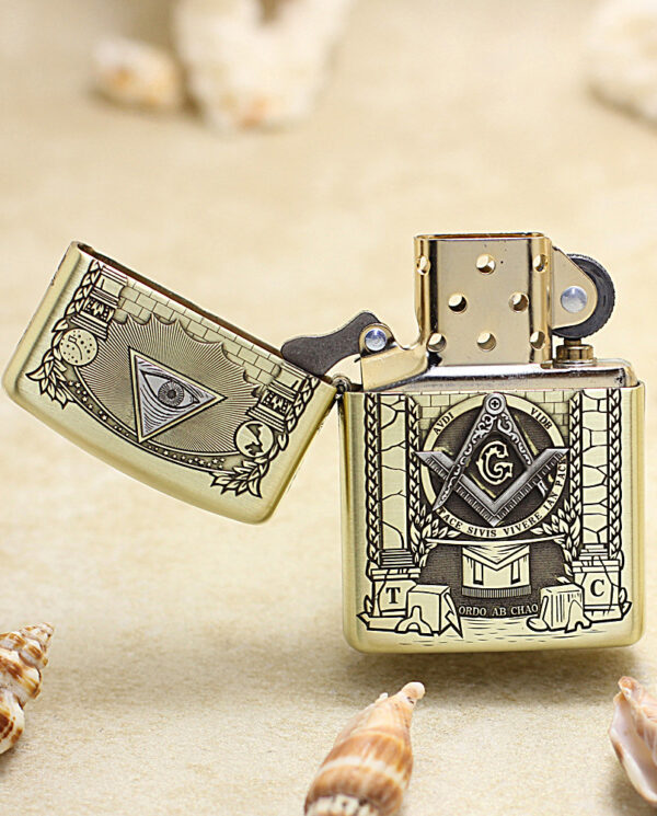 Gold Zippo lighter with Freemason square-and-compass emblem and open lid, set on a sandy beach with seashells nearby.