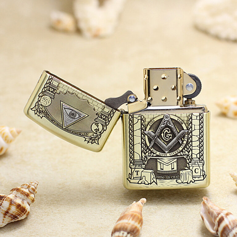 Gold Zippo lighter with Freemason square-and-compass emblem and open lid, set on a sandy beach with seashells nearby.