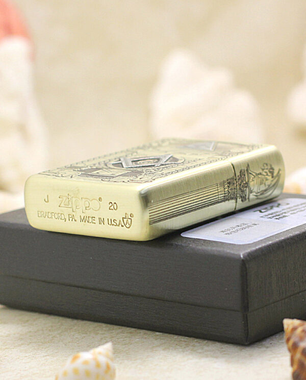 Brass engraved Zippo lighter resting on a black gift box with a blurred seashell backdrop.