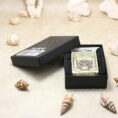Gold-tone Zippo lighter with engraved design in a black presentation box on a sandy surface surrounded by seashells.