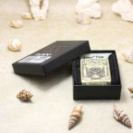 Gold-tone Zippo lighter with engraved design in a black presentation box on a sandy surface surrounded by seashells.