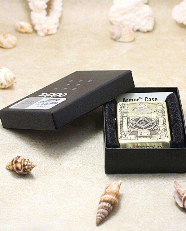 Gold-tone Zippo lighter with engraved design in a black presentation box on a sandy surface surrounded by seashells.