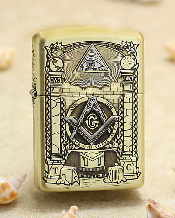 Gold Zippo lighter with Masonic symbols (eye in triangle, compass and square with G) displayed on a sandy beach with seashells nearby.