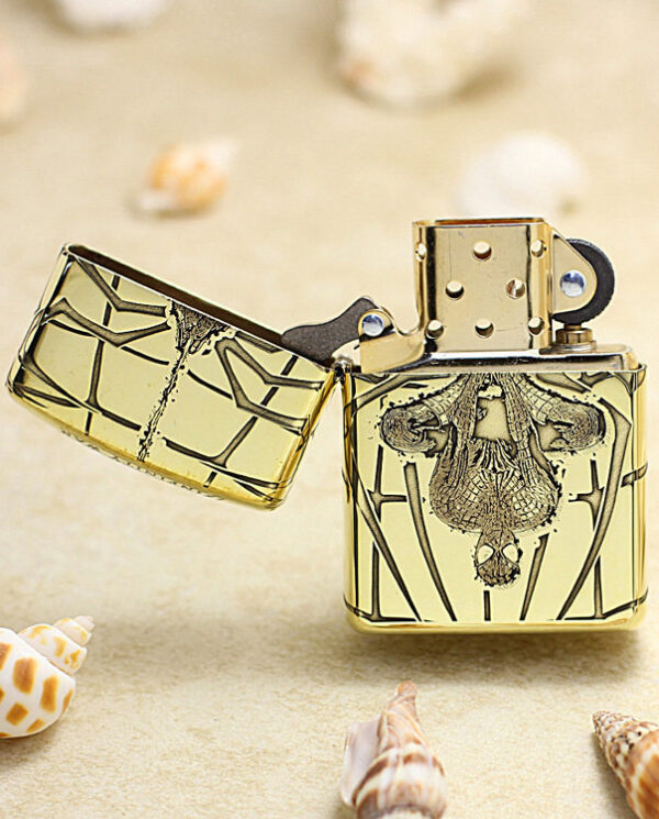 Gold engraved Zippo lighter with ornate pattern, open on a sandy beach with seashells nearby.
