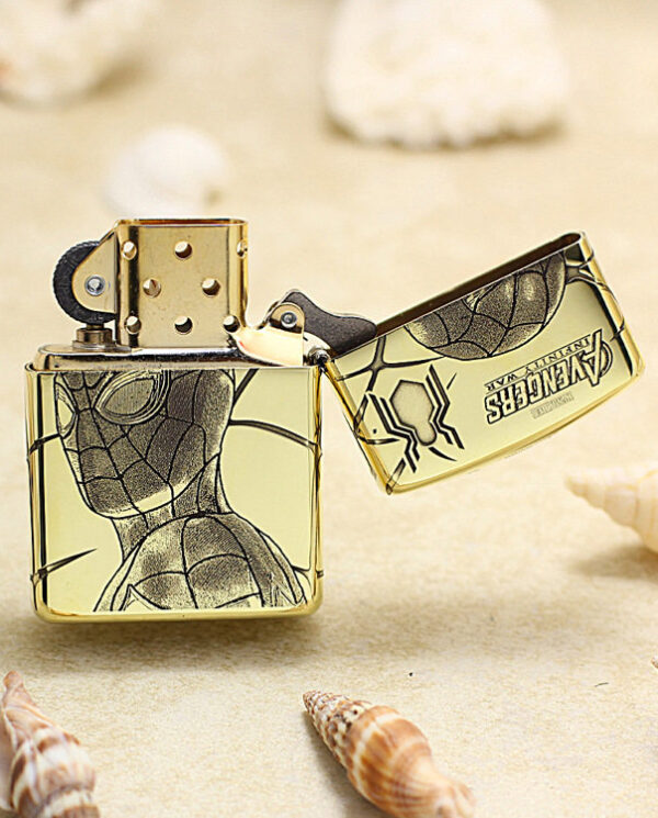 Gold-tone Zippo-style lighter with Spider-Man web design, open lid on a sandy beach with seashells nearby.