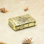 Gold metal lighter with an engraved design rests on sandy surface near small seashells. A close-up of a gold lighter on a beach scene with shells nearby. A gold engraved lighter positioned on beige sand with scattered shells. Shiny gold pocket lighter with etched artwork lying on a beach with shells. Engraved gold lighter on sand among tiny sea shells, foreground focus.