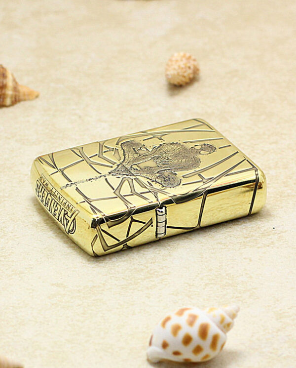 Gold metal lighter with an engraved design rests on sandy surface near small seashells. A close-up of a gold lighter on a beach scene with shells nearby. A gold engraved lighter positioned on beige sand with scattered shells. Shiny gold pocket lighter with etched artwork lying on a beach with shells. Engraved gold lighter on sand among tiny sea shells, foreground focus.