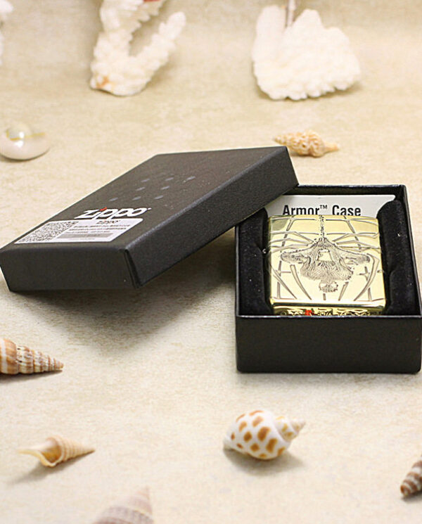 Gold Zippo lighter in a black gift box on beige sand, engraved with an insect, surrounded by seashells on a beach backdrop.