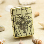 Gold-toned lighter with an ornate skull and geometric engraving, set on a sandy beach with seashells nearby.
