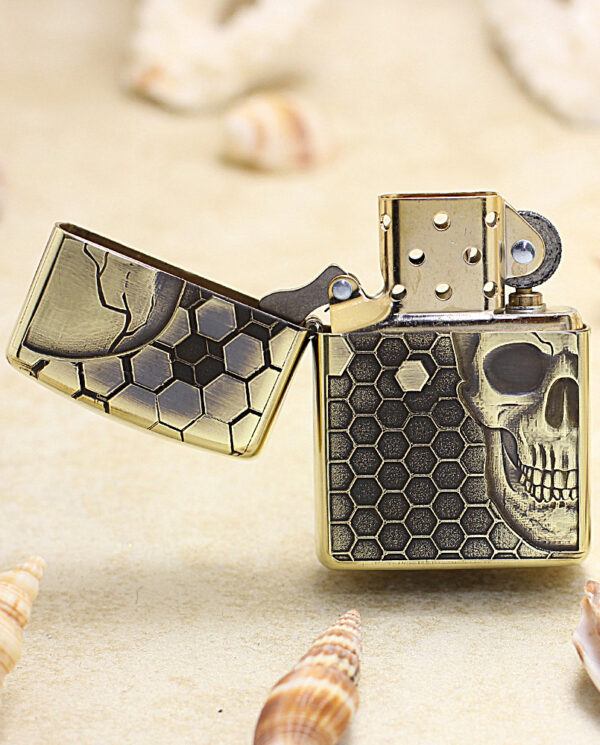 KZ240-02 Gold-toned Zippo-style lighter with a skull and honeycomb pattern, lid open, resting on sandy surface with seashells nearby