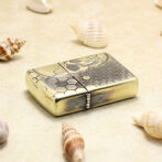 Gold-toned lighter with a skull and honeycomb pattern lying on sand among seashells.