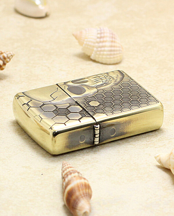 KZ240-04 Gold-toned lighter with a skull and honeycomb pattern lying on sand among seashells.