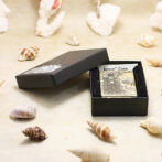 Metal lighter with a skull and honeycomb pattern in an open black gift box on a sandy surface with seashells nearby.