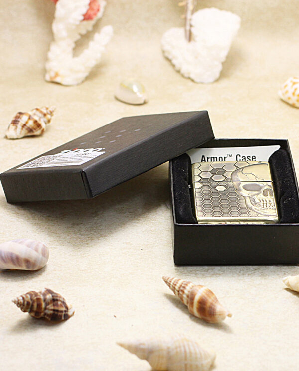 KZ240-06 Metal lighter with a skull and honeycomb pattern in an open black gift box on a sandy surface with seashells nearby.