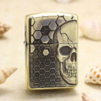 Gold-tone lighter with a skull motif over a hexagonal honeycomb pattern, standing upright on a sandy surface.