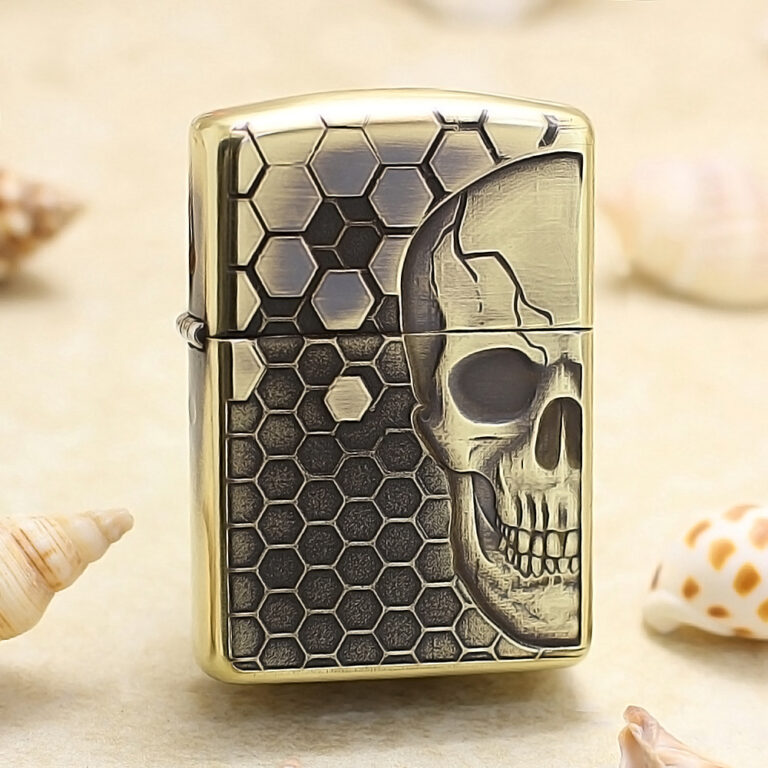 Gold-tone lighter with a skull motif over a hexagonal honeycomb pattern, standing upright on a sandy surface.