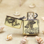 Gold Zippo lighter with Avengers artwork, open to show the flint wheel, resting on sand with seashells nearby.