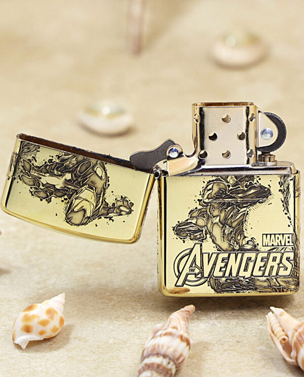 KZ241-02 Gold Zippo lighter with Avengers artwork, open to show the flint wheel, resting on sand with seashells nearby.