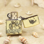 Gold Avengers-themed Zippo lighter, open, resting on sand with seashells nearby.