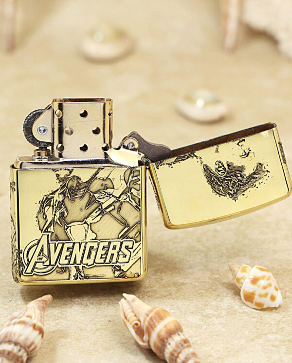 KZ241-03 Gold Avengers-themed Zippo lighter, open, resting on sand with seashells nearby.