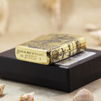 Close-up of a polished gold Zippo lighter with engraved artwork resting on a black box, seaside shells in the foreground