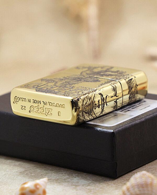 KZ241-05 Close-up of a polished gold Zippo lighter with engraved artwork resting on a black box, seaside shells in the foreground