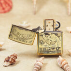 Brass Zippo lighter open to reveal a camel engraving and the text "EST. 1913 TURKISH AND DOMESTIC BLEND" on a sandy beach with seashells nearby.