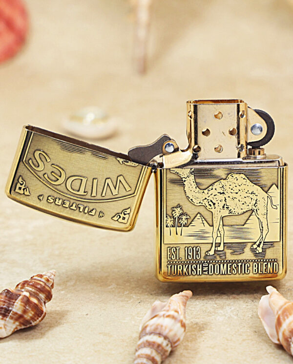 KZ242-02 Brass Zippo lighter open to reveal a camel engraving and the text "EST. 1913 TURKISH AND DOMESTIC BLEND" on a sandy beach with seashells nearby.