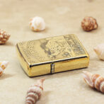 Gold-tone Zippo lighter with engraved map on lid, resting on sandy surface among seashells