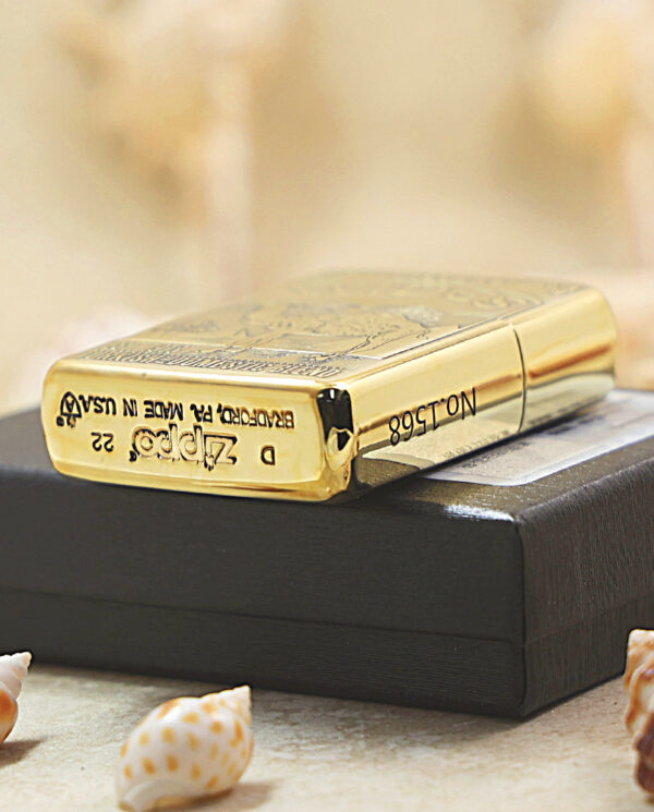 KZ242-05 Gold Zippo lighter with engraved design resting on a black box, surrounded by seashells on a sandy surface behind.