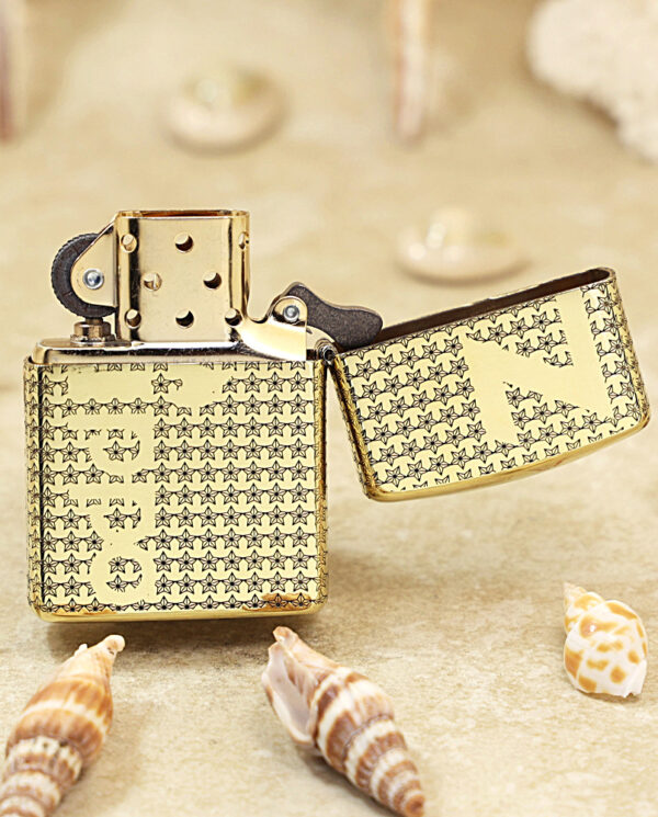 Gold patterned lighter on sand with its lid open, surrounded by seashells on a beach.