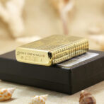 Gold patterned Zippo lighter resting on a black box, with seashells in the foreground.