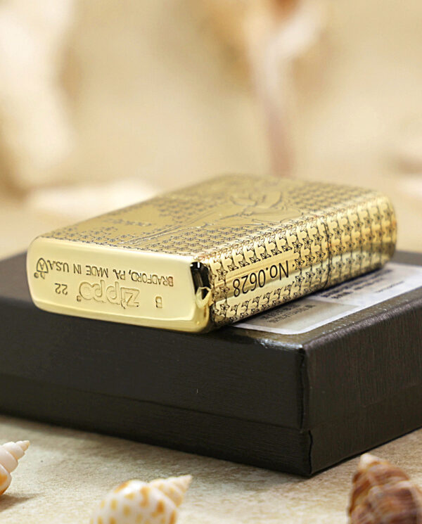 Gold patterned Zippo lighter resting on a black box, with seashells in the foreground.