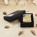Gold engraved Zippo lighter in an open black presentation box on a sandy surface with seashells around it.